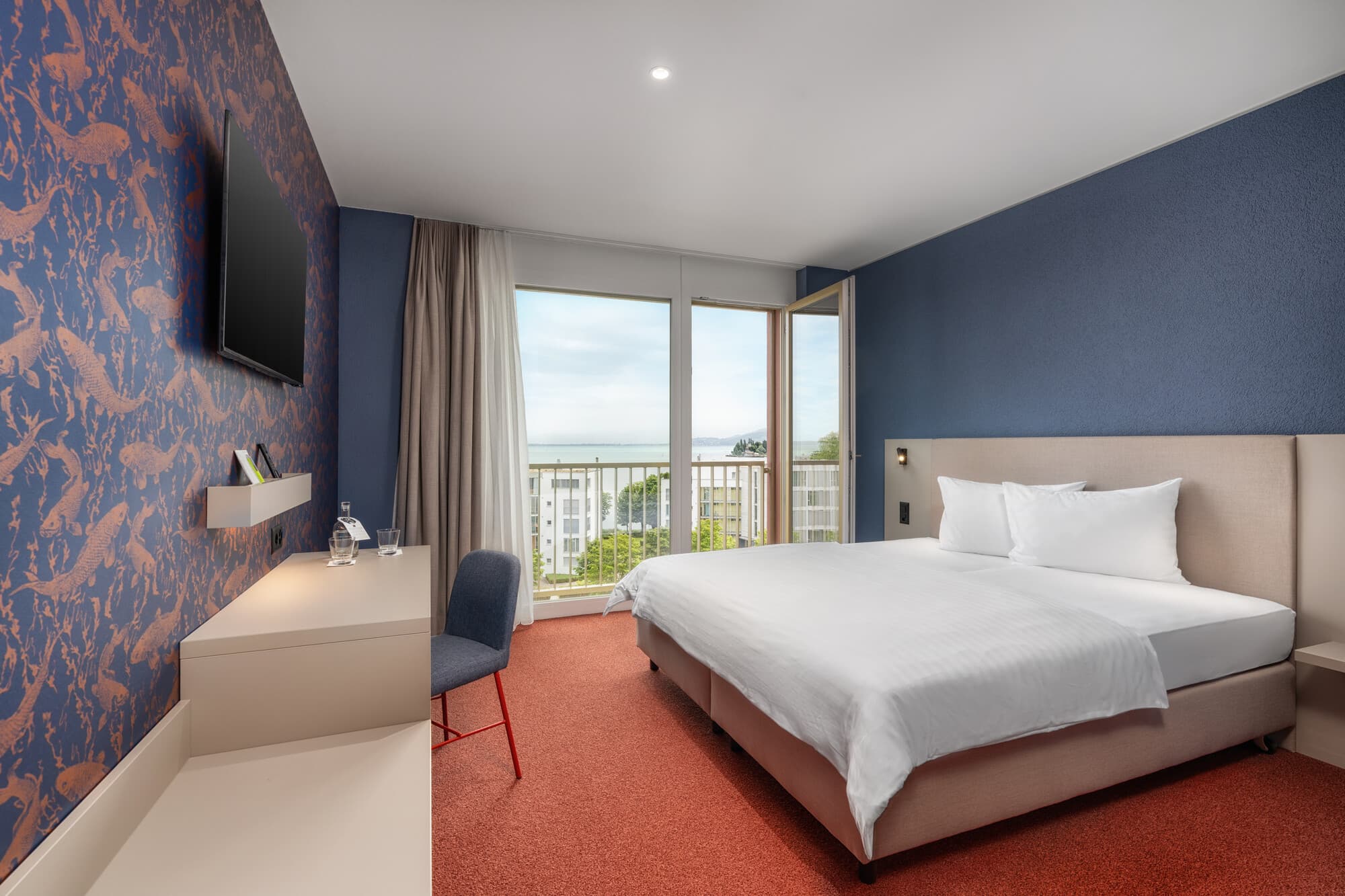 A deluxe double room in Arbon with blue wall paint, modern furnishings, red carpeted floor, and a view of the lake from the window.