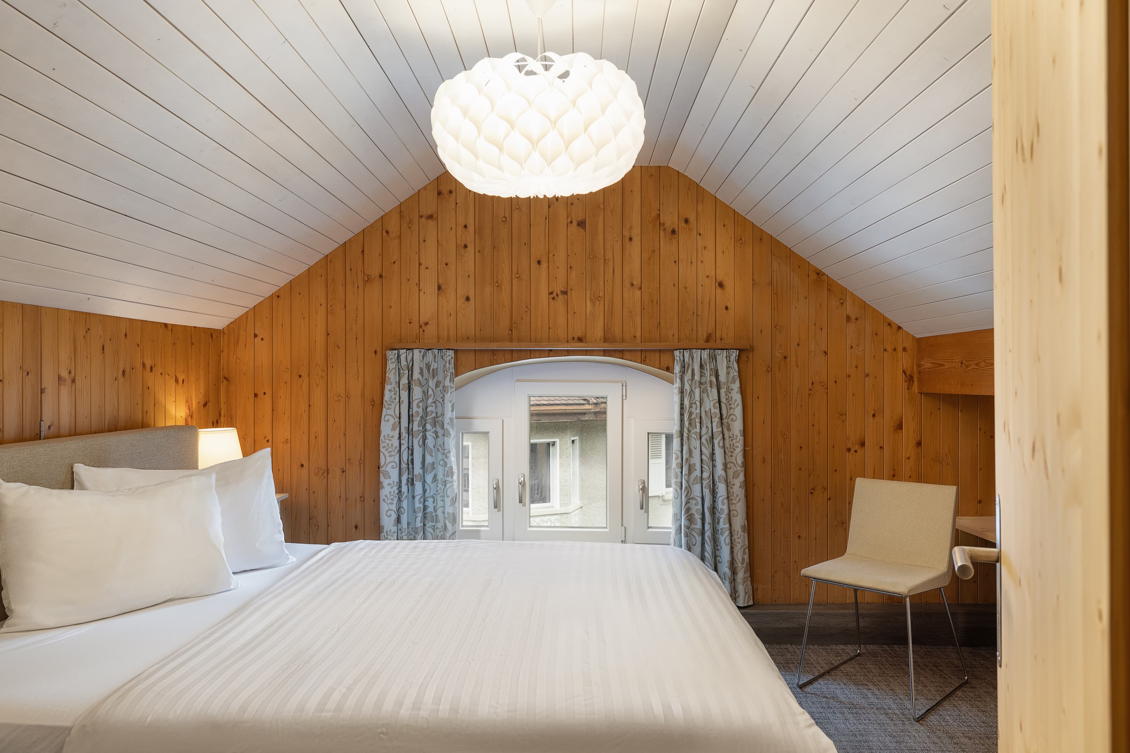 A bright superior apartment room in the Hotel Krone with wood paneling, a large double bed, a modern chair, and a stylish chandelier.
