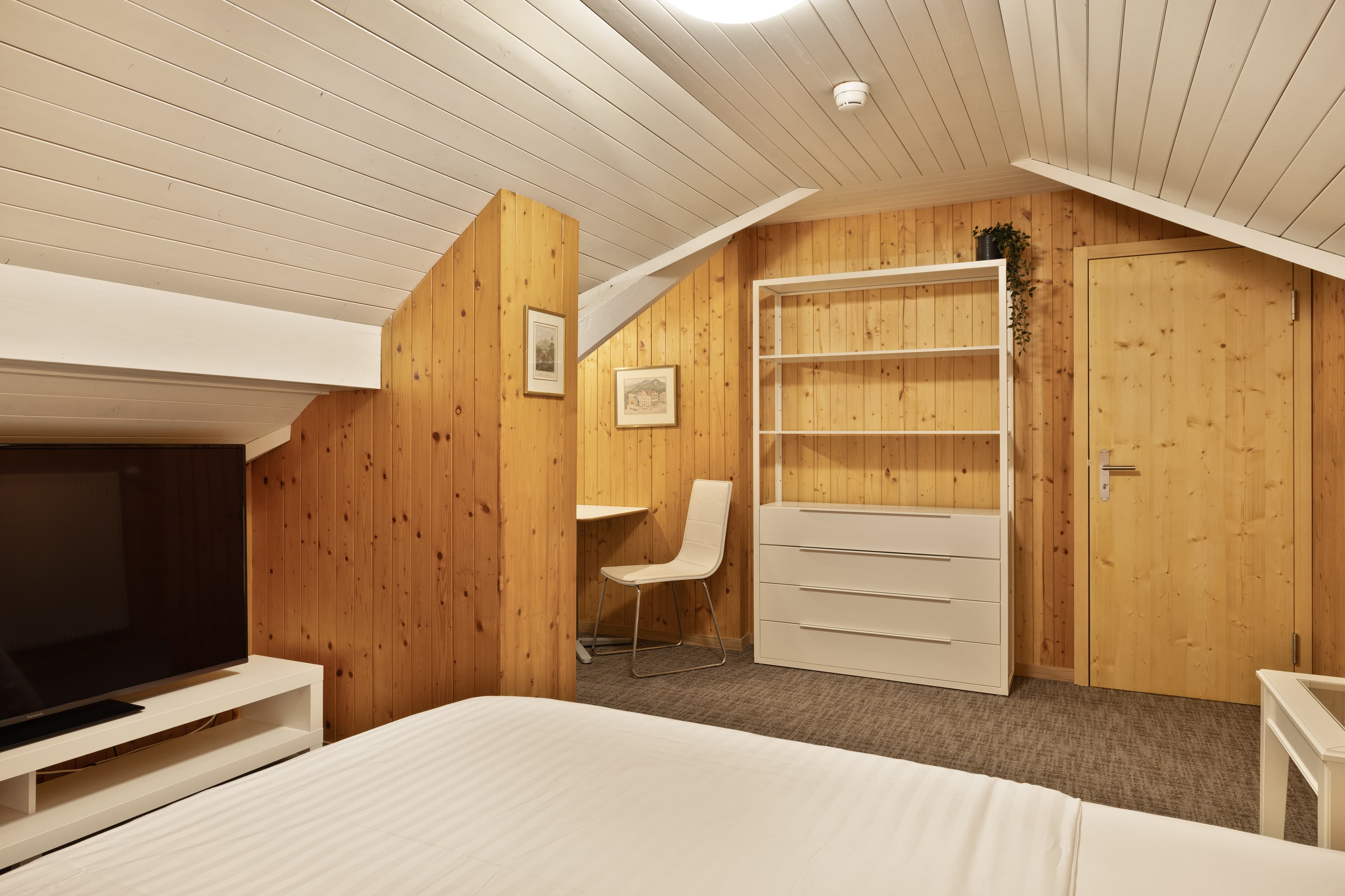 A cozy hotel room with wood paneling a bed a TV white furniture and a small desk with a chair