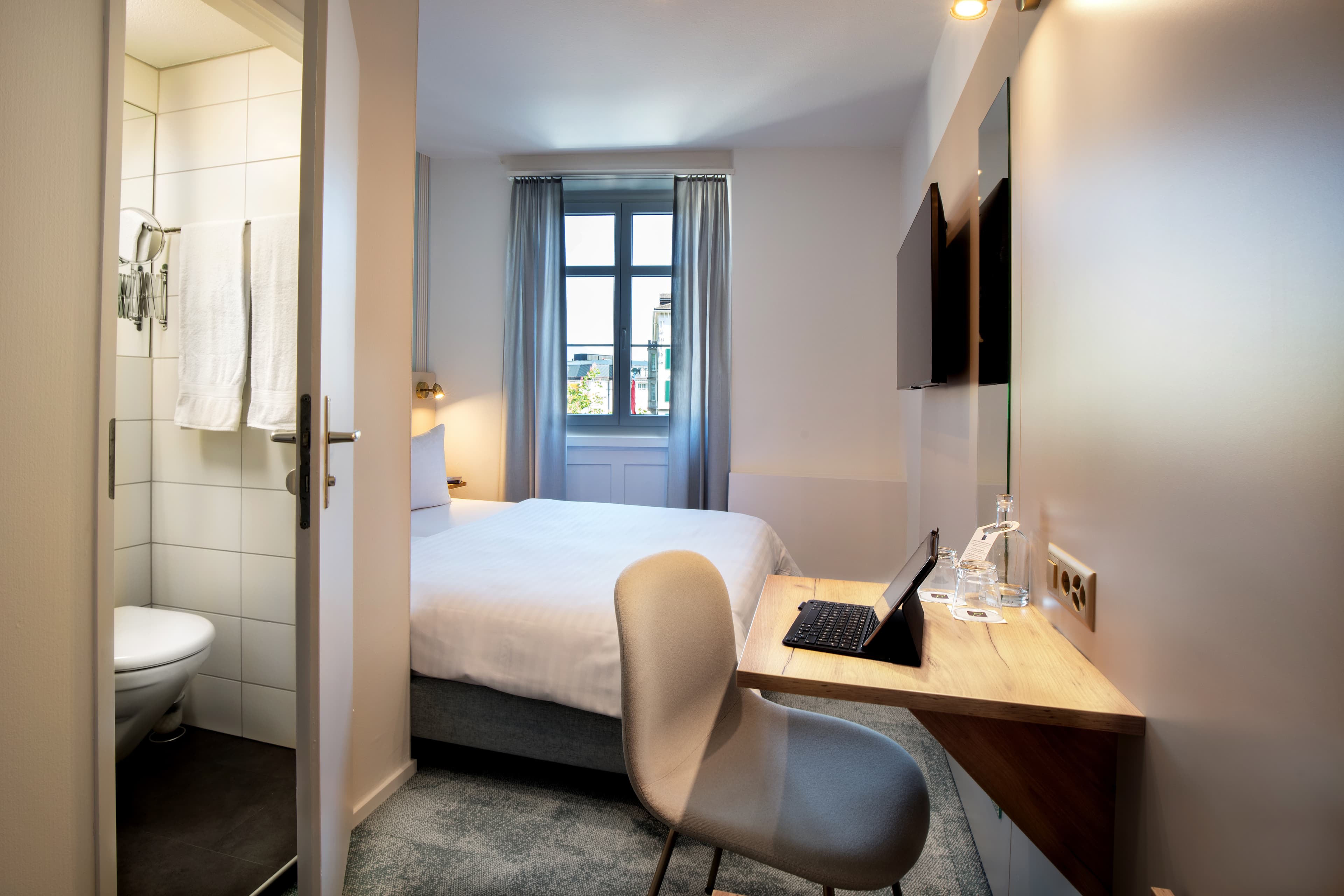 A standard double room at Hotel Krone by b smart with a bed a desk and an adjoining bathroom