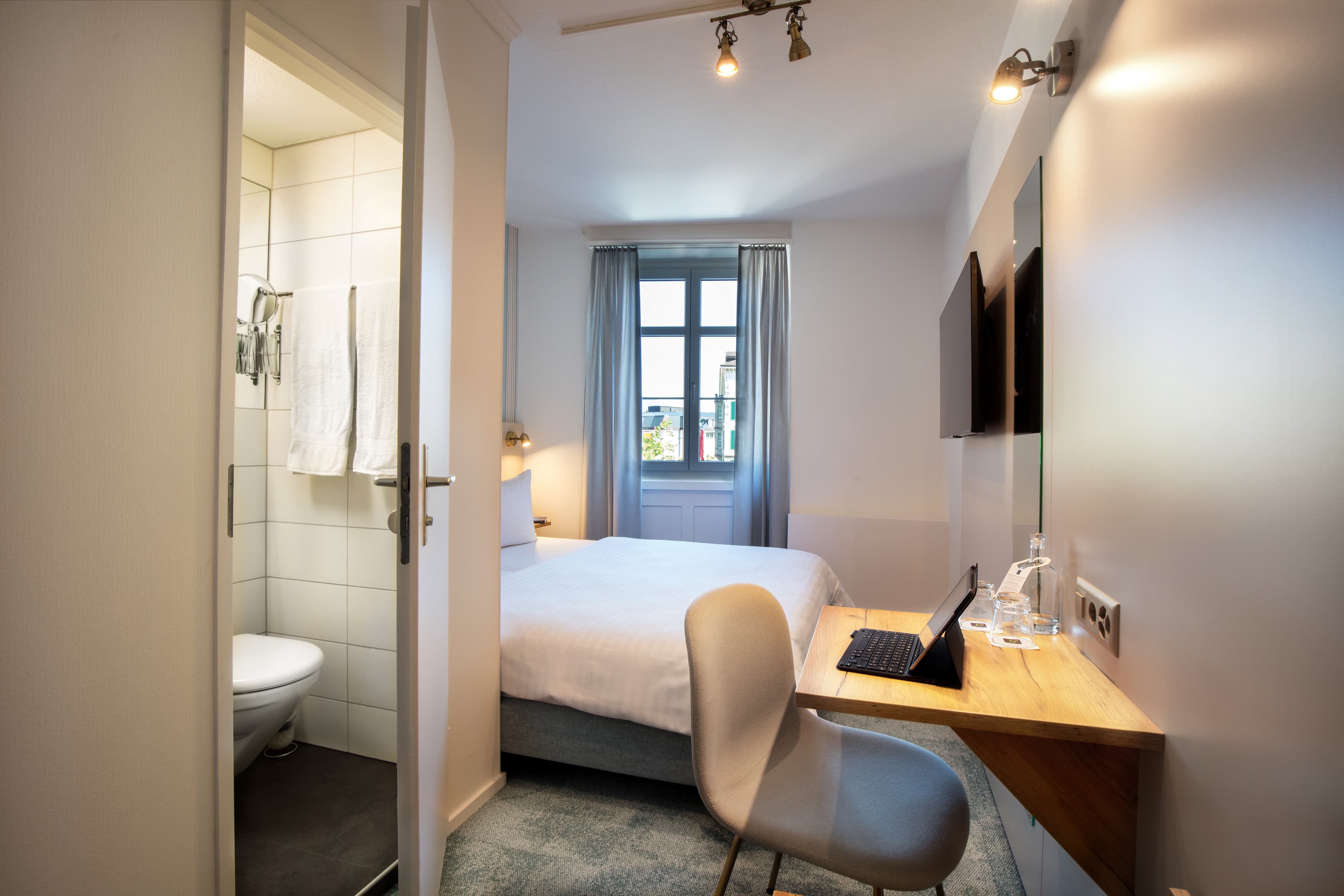 A modern double room at Hotel Krone by b_smart with a desk, chair, an en-suite bathroom, and a window with blackout curtains