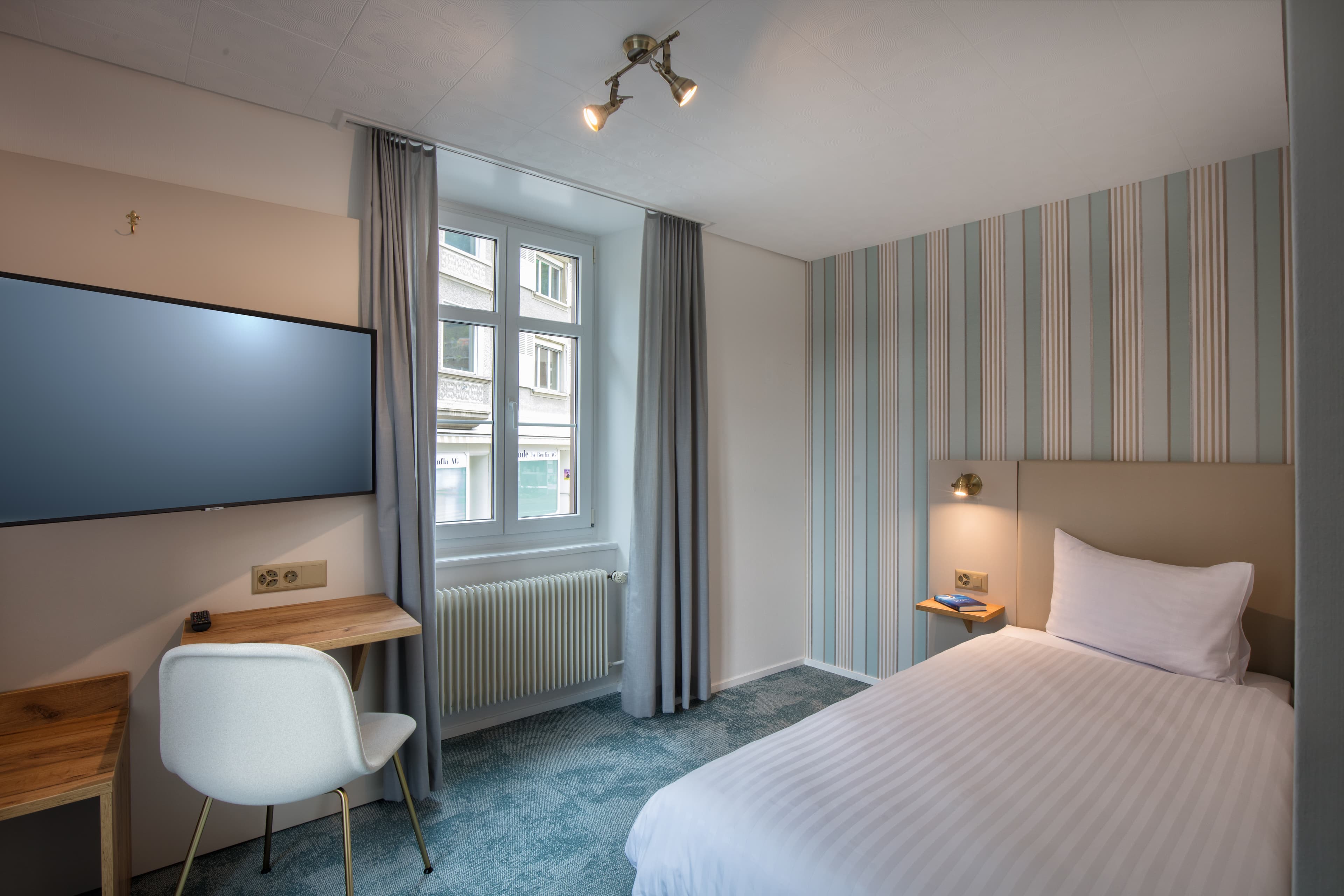 A single room at Hotel Krone by b_smart with a bed a desk a television a window and understated striped wallpaper